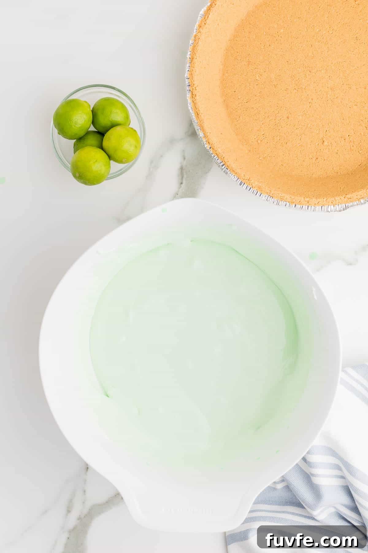 No-Bake Key Lime Jello Pie with Cool Whip Topping 6 Smooth key lime pie filling in a mixing bowl before being poured into the crust. Fresh limes and a prepared graham cracker crust are positioned beside the bowl.