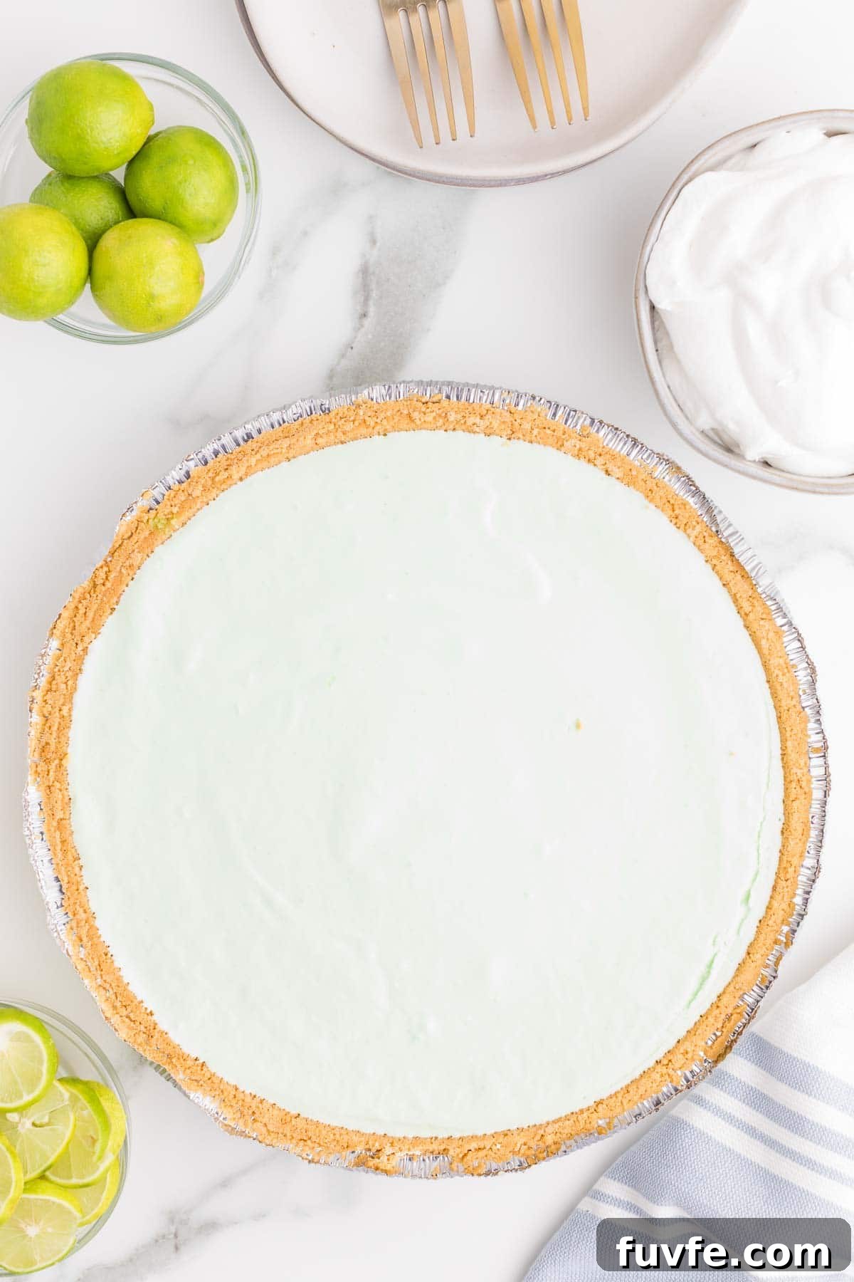 No-Bake Key Lime Jello Pie with Cool Whip Topping 7 Whole key lime Jello pie set in a graham cracker crust before slicing. Fresh limes and whipped topping are arranged nearby as serving accompaniments.