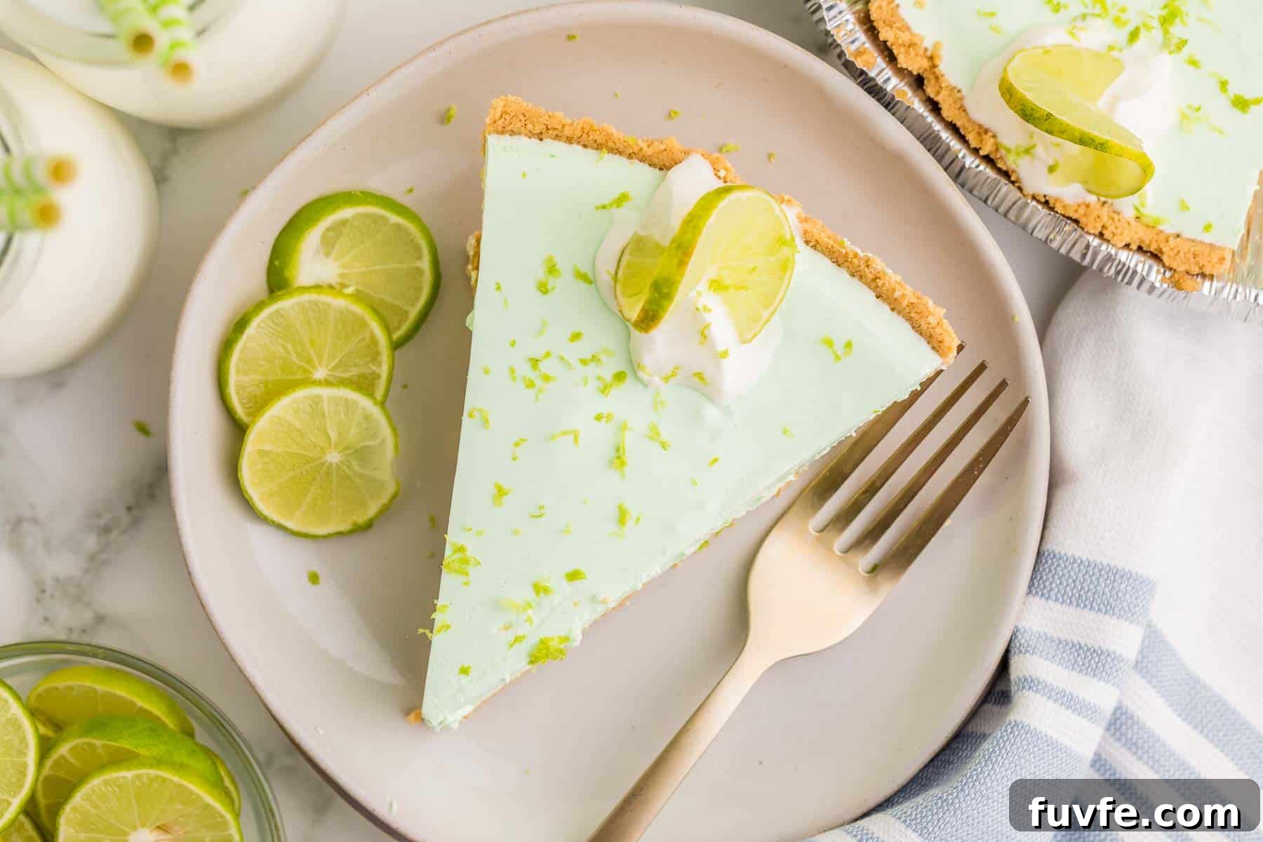 No-Bake Key Lime Jello Pie with Cool Whip Topping 10 Overhead view of a slice of key lime Jello pie topped with whipped cream, lime zest, and a lime slice. Fresh lime slices and a fork sit beside the plate.