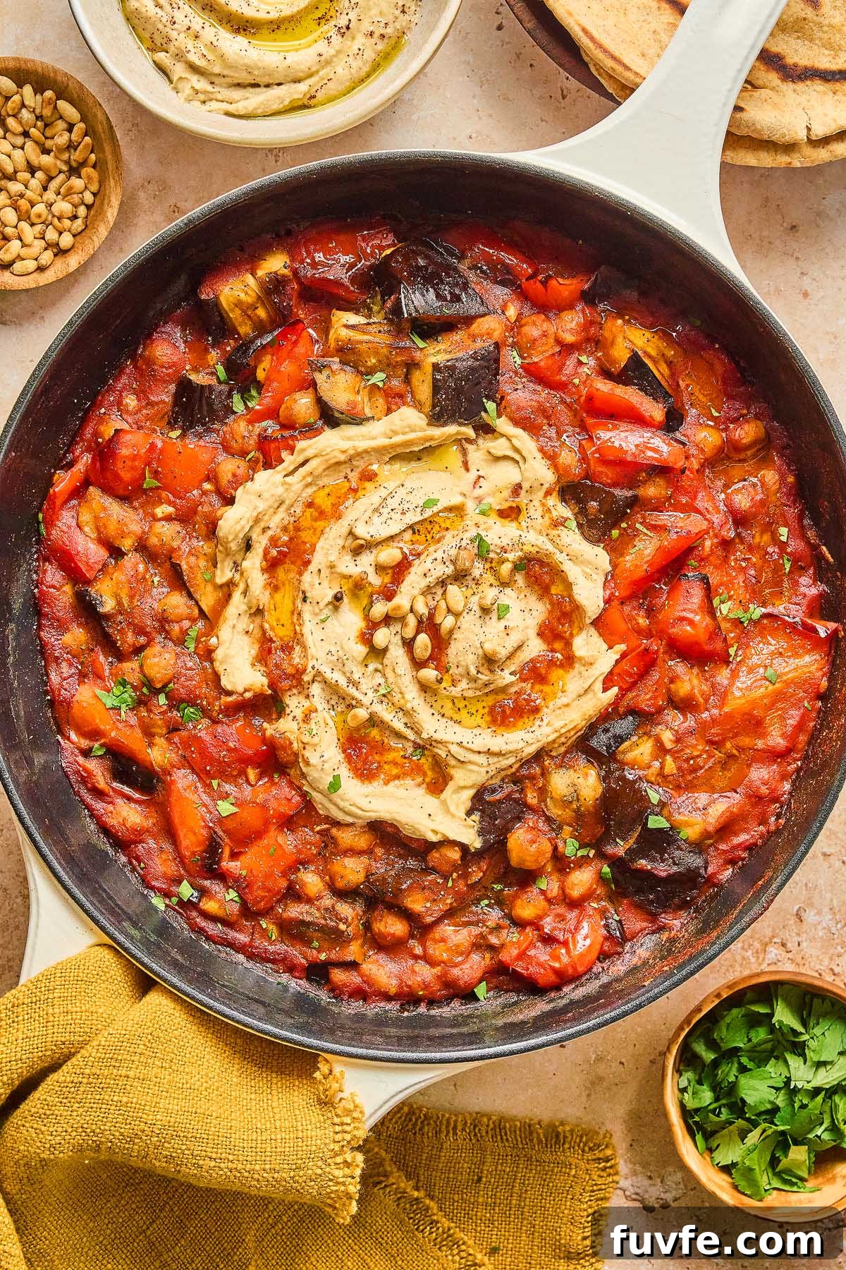 One-Pan Mediterranean Chickpea Skillet with Tomatoes & Spinach 10 A Mediterranean chickpea skillet brimming with a tomato-based stew, elegantly topped with a swirl of hummus and pine nuts. Encircling the skillet are small bowls filled with extra pine nuts, parsley, and hummus for added layers of flavor.