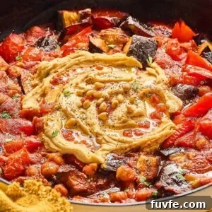 One-Pan Mediterranean Chickpea Skillet with Tomatoes & Spinach 13 A pan of shakshuka with a swirl of hummus and sprinkled pine nuts in the center, reminiscent of a Mediterranean chickpea skillet. Surroundings include tomatoes, eggplant, bell peppers, and herbs.