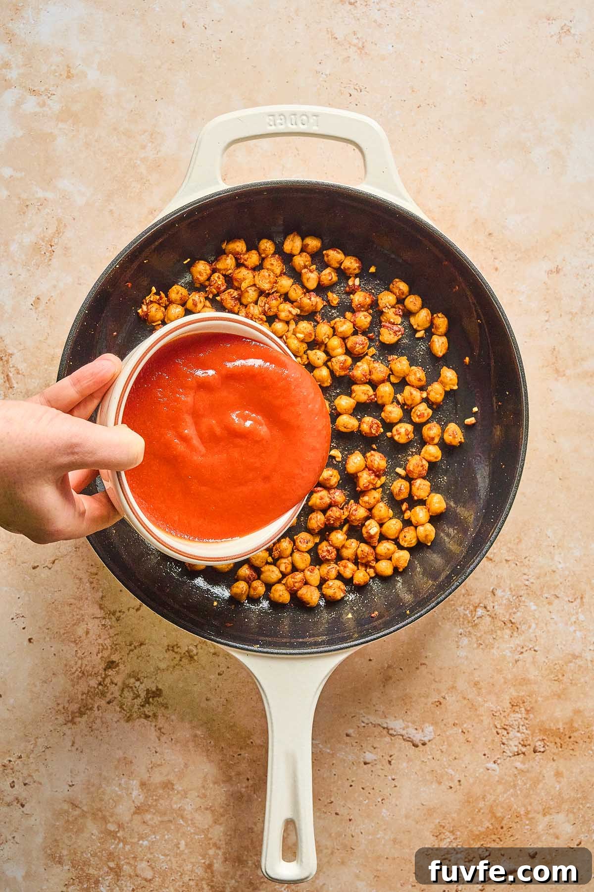 One-Pan Mediterranean Chickpea Skillet with Tomatoes & Spinach 17 A hand effortlessly pours tomato sauce from a pristine white bowl onto a delightful Mediterranean chickpea skillet, sizzling gently in a white pan.