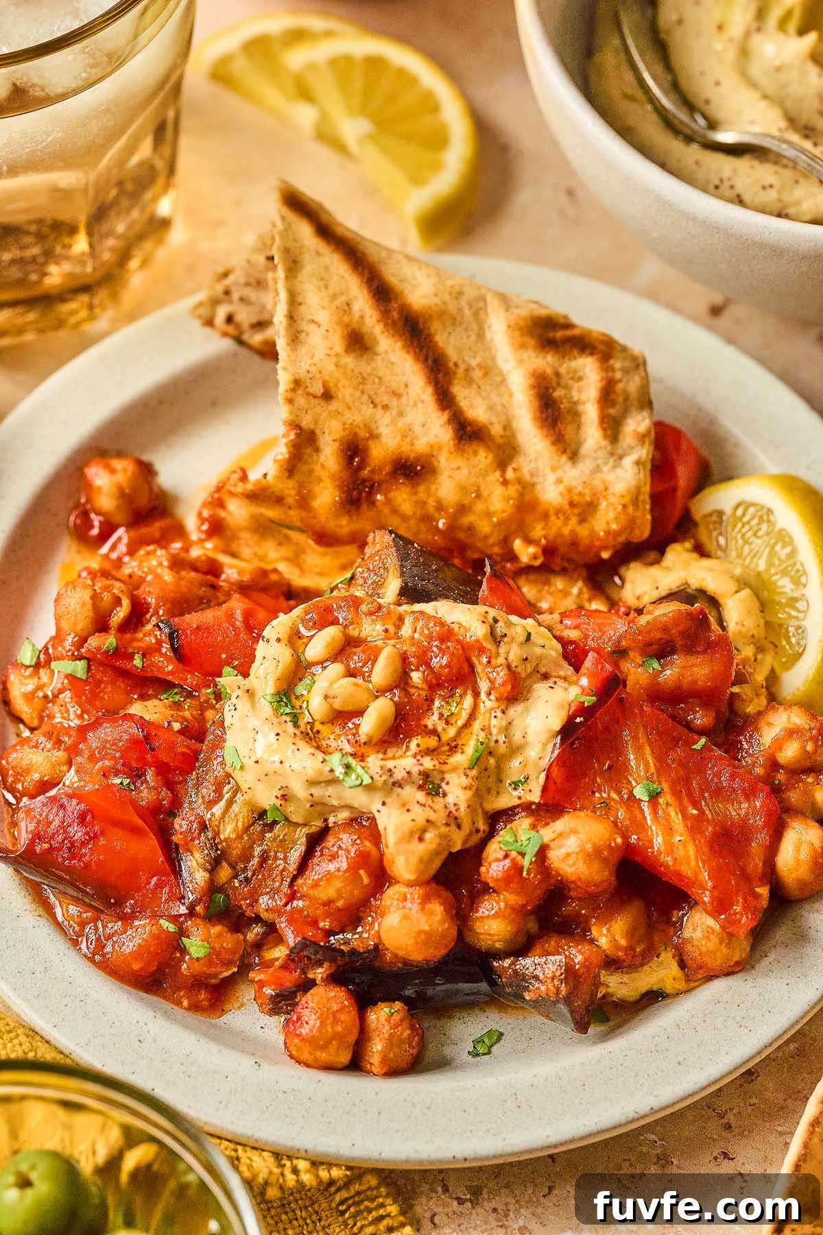 One-Pan Mediterranean Chickpea Skillet with Tomatoes & Spinach 5 A Mediterranean chickpea skillet featuring roasted vegetables and hummus, garnished with pine nuts and served alongside grilled flatbread and a lemon wedge.