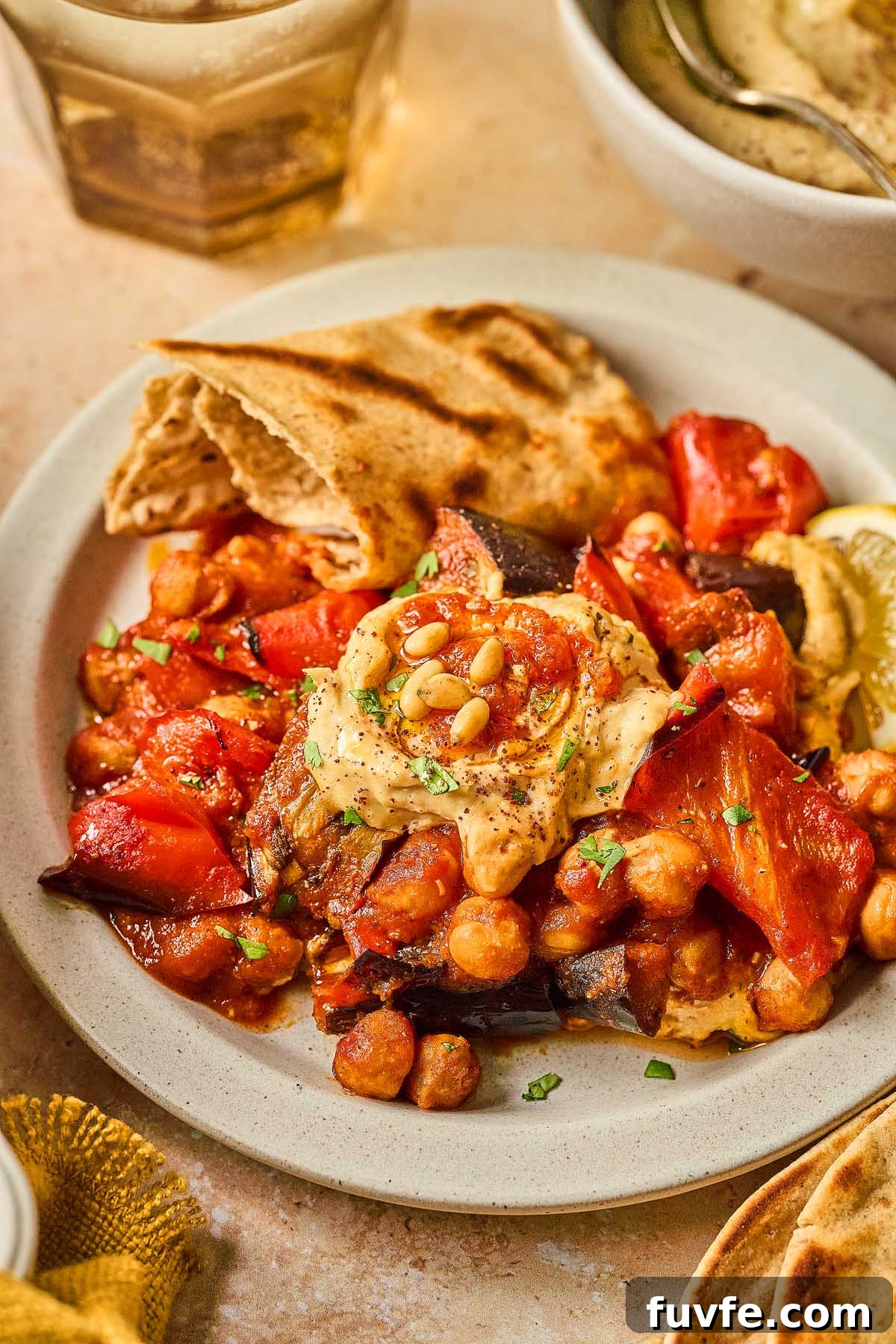 One-Pan Mediterranean Chickpea Skillet with Tomatoes & Spinach 8 A Mediterranean chickpea skillet features a plate of roasted vegetables and chickpeas in tomato sauce, topped with pine nuts and served alongside pita bread. A refreshing glass of liquid and a bowl of dip await in the background, enhancing this vibrant culinary experience.