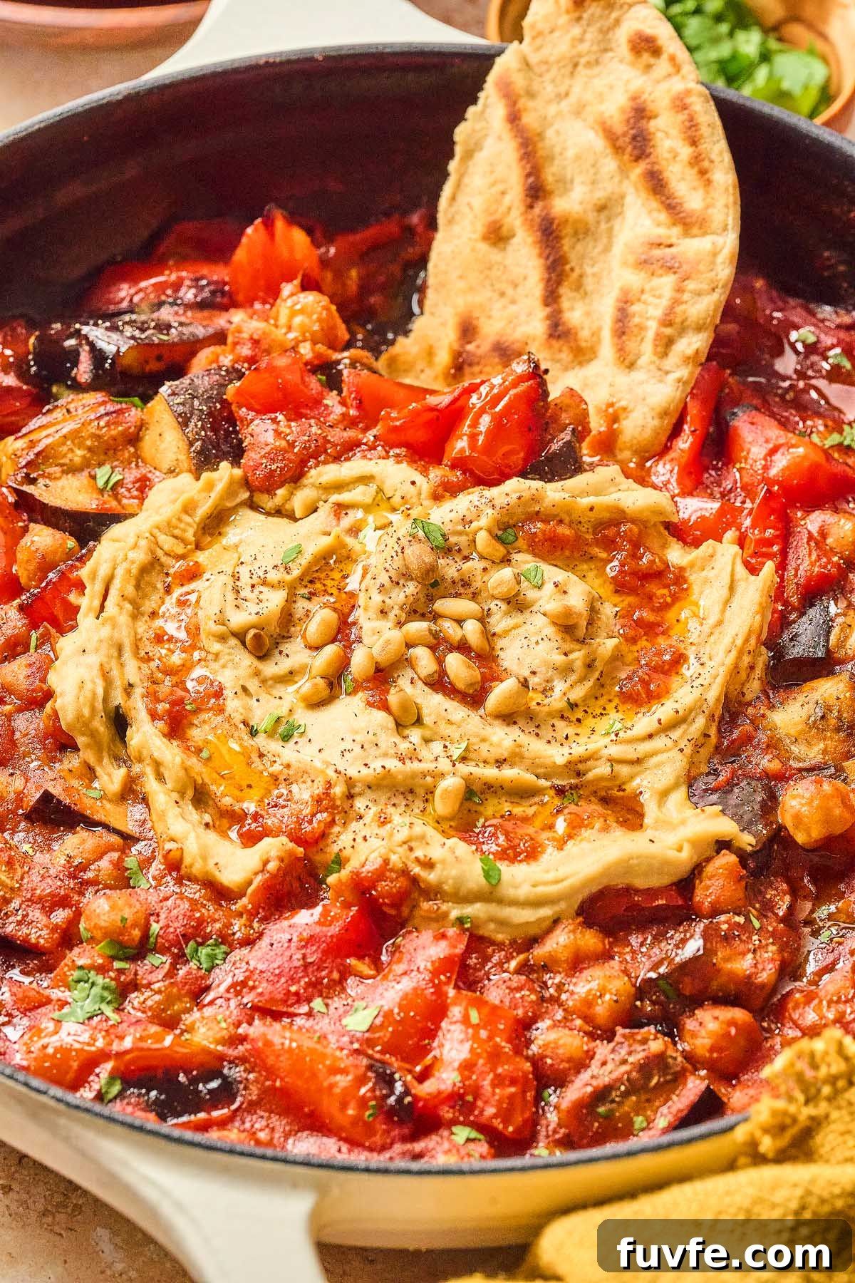 One-Pan Mediterranean Chickpea Skillet with Tomatoes & Spinach 9 A skillet filled with a chunky Mediterranean chickpea and tomato vegetable stew, topped with a swirl of hummus, pine nuts, and herbs. A piece of bread is dipped in the mixture.