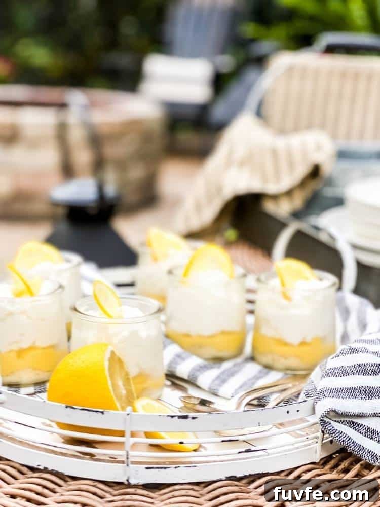 Six small clear glass jars of Lemon Bar in a Jar sitting on a white wood and metal serving tray with spoons and a striped tea towel on a wicker table at an outdoor picnic.