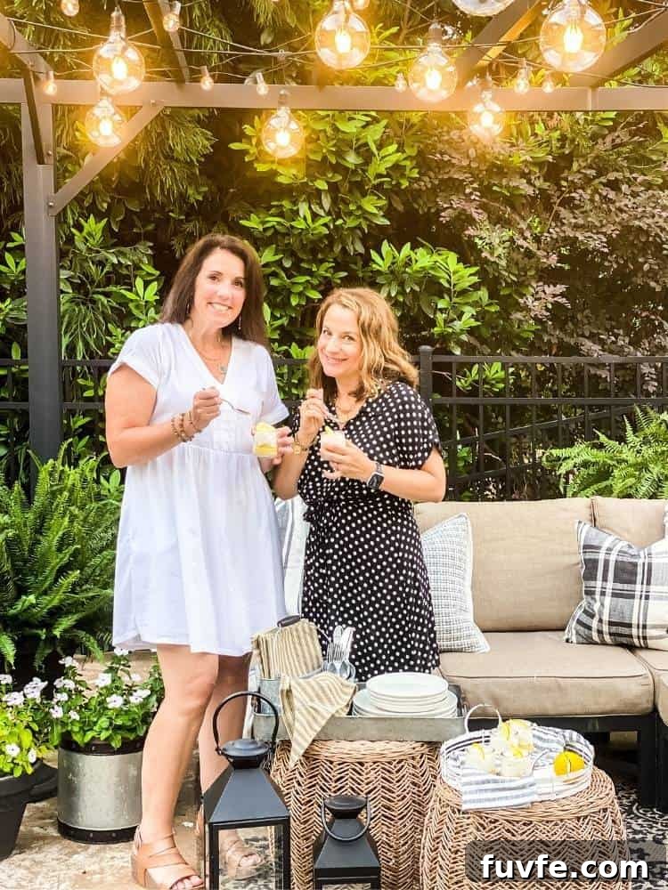 Two women holding cups of lemon bars in a jar standing under a pergola strung with different sizes of string lights. The background is all kinds of shrubs. A tan outdoor sofa sits under the pergola with wicker tables, lanterns, and potted plants.