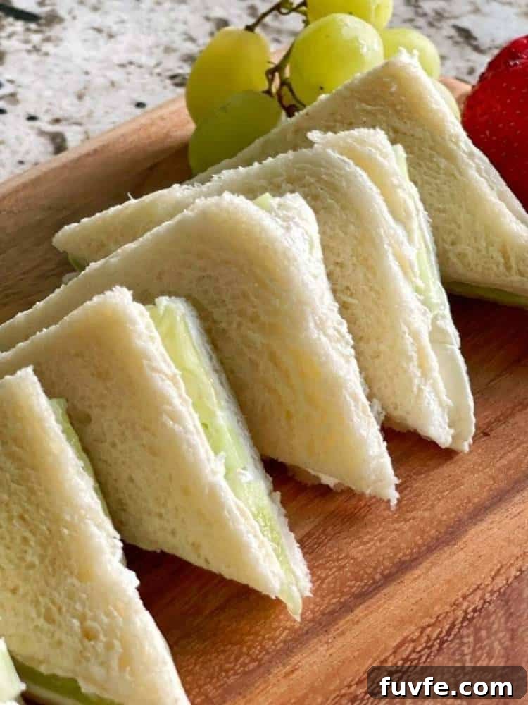 Wood cutting board with 5 cucumber sandwiches on white bread cut in triangles with the crusts removed. Fresh grapes and strawberries garnish the board