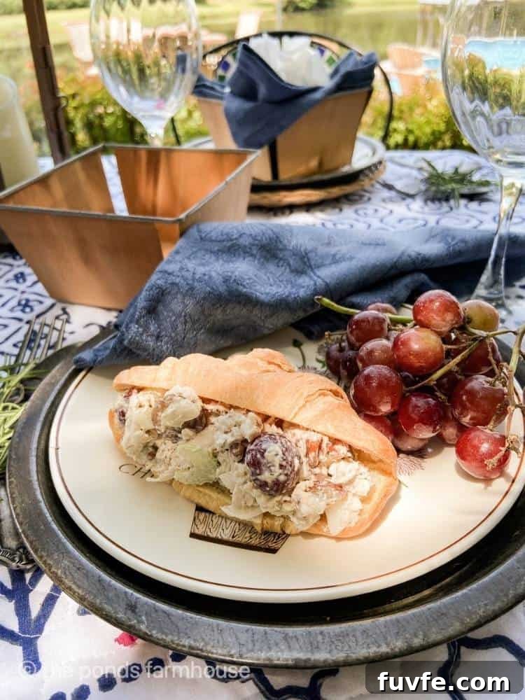 A croissant stuffed with chicken salad sits on a plate with a bunch of red grapes. A blue cloth napkin is arranged at the top of the plate and wine glasses sit on the table with small wooden baskets.