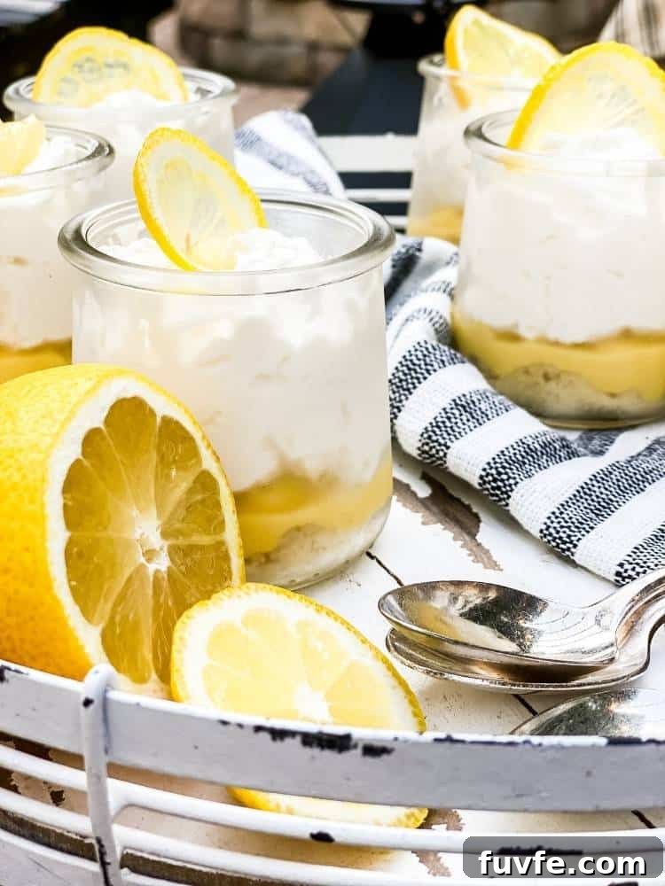 Five small clear glass jars of Lemon Bar in a Jar sitting on a white wood and metal serving tray with spoons and a striped tea towel. A cut lemon is the garnish.