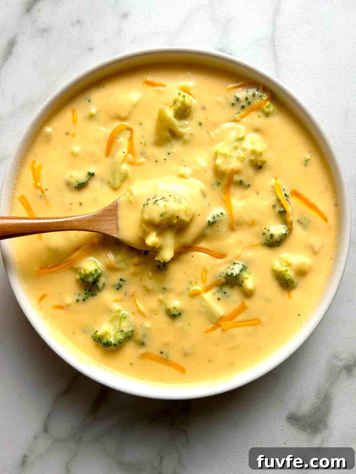overhead shot of broccoli cheddar soup in white bowl; wooden spoon lifting up some of the soup.