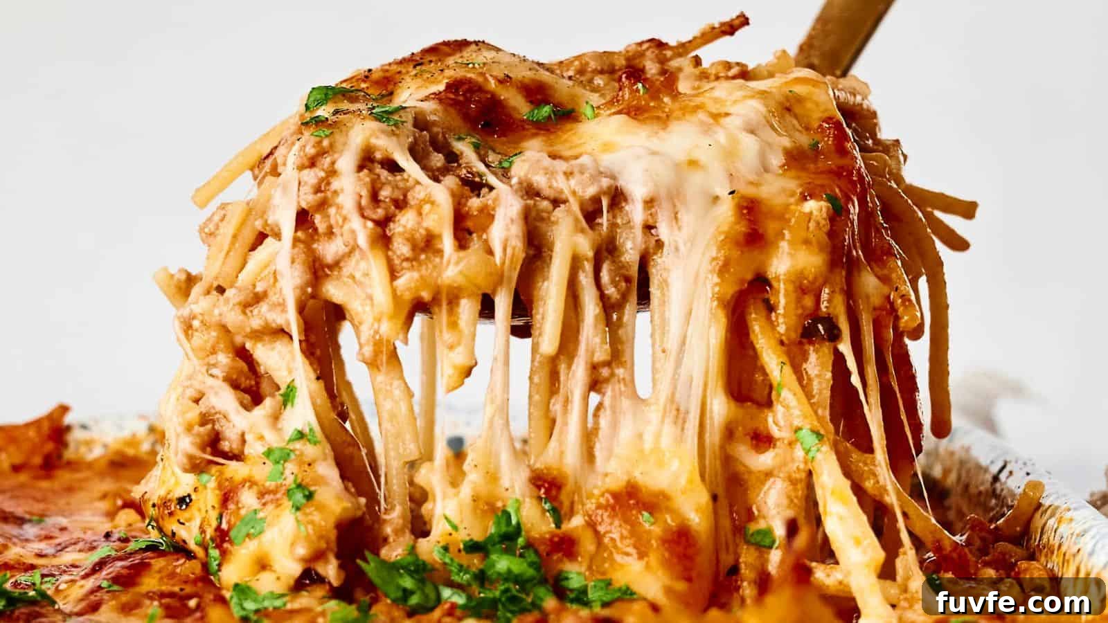 A serving of cheesy baked spaghetti is being lifted with a utensil. The dish is topped with melted cheese and garnished with herbs.