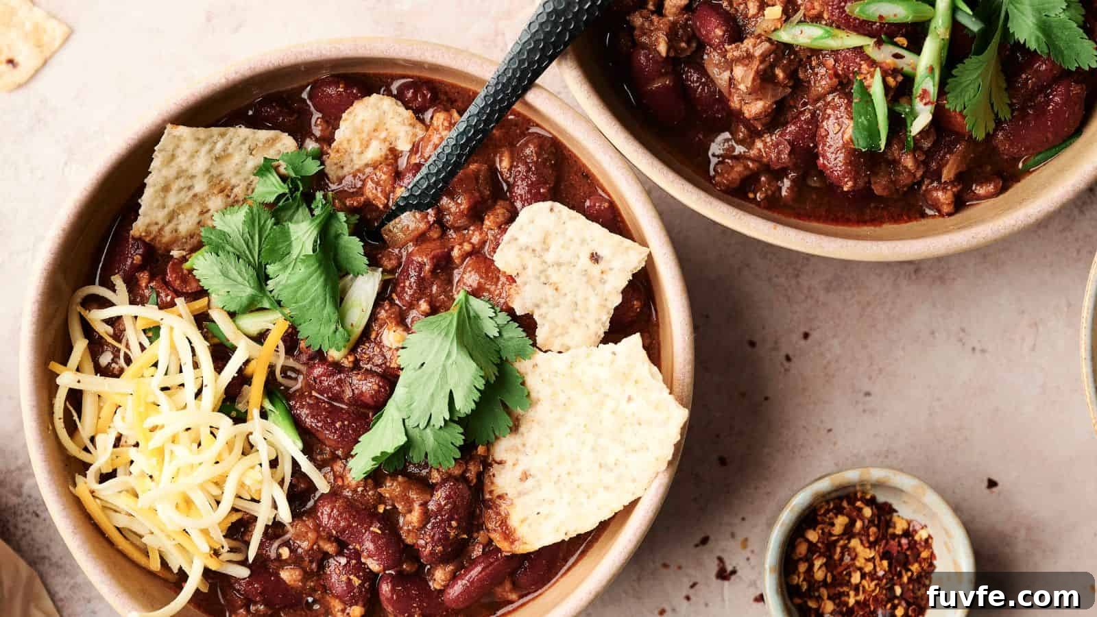 Two bowls of chili with kidney beans, meat, tortilla chips, shredded cheese, and cilantro. A black spoon in one bowl. Additional chili flakes in a small dish nearby.