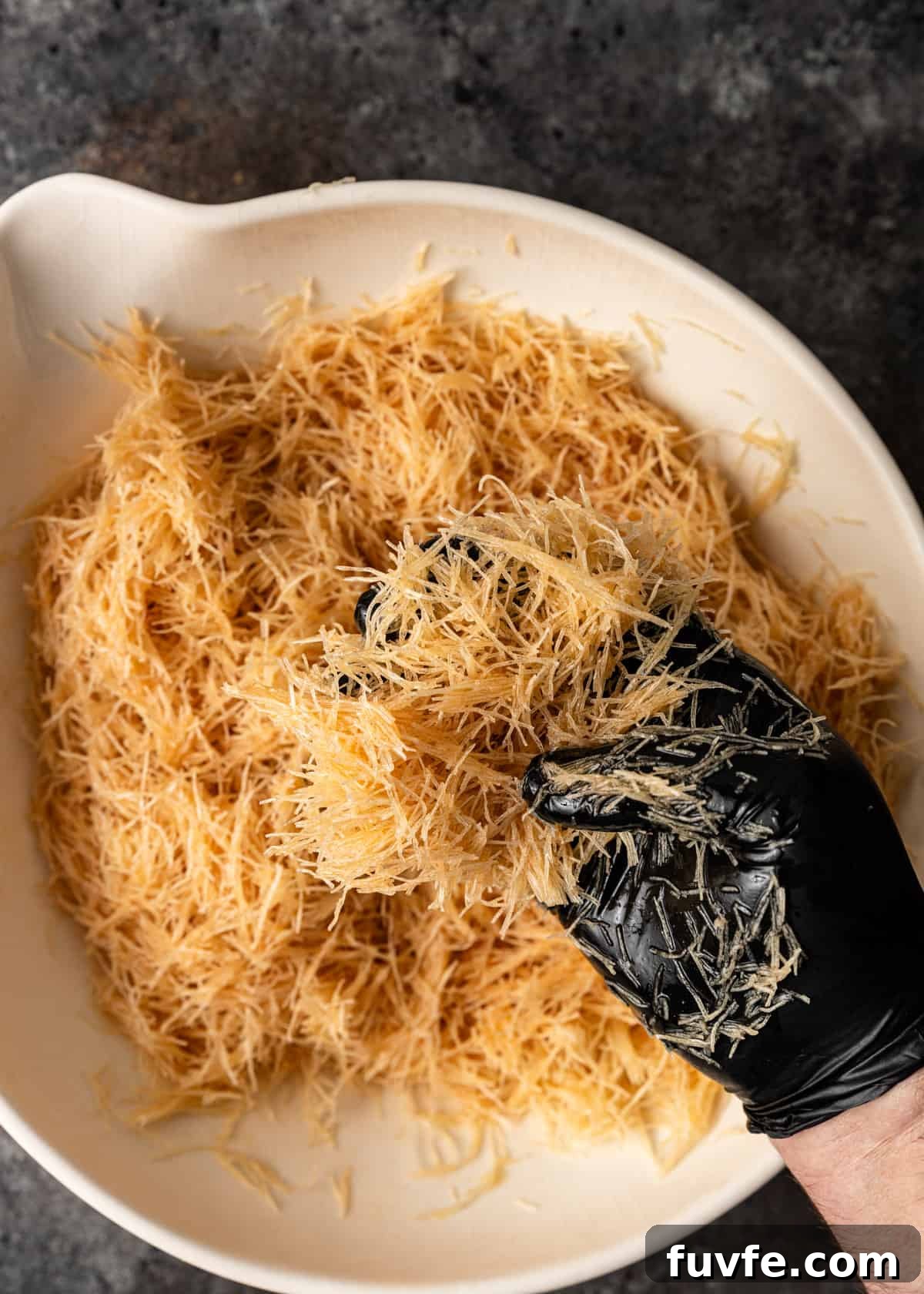 overhead: a black gloved hand mixing shredded filo dough with melted ghee