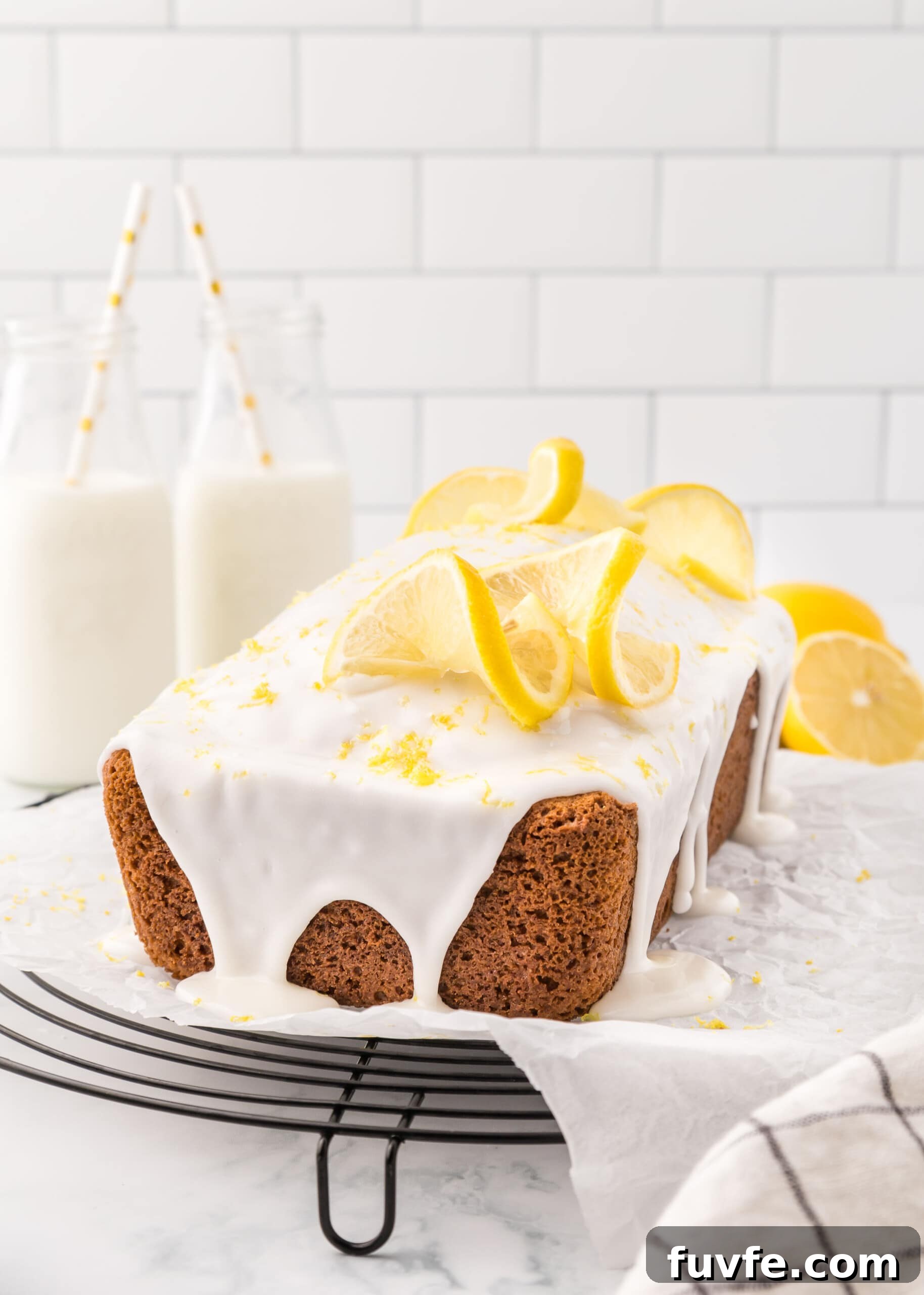 Zesty Iced Lemon Loaf Recipe for a Moist Citrus Cake 2 Lemon loaf with icing sprinkled with lemon zest and topped with two twisted lemon slices accented with glass milk bottles with white and yellow straws.