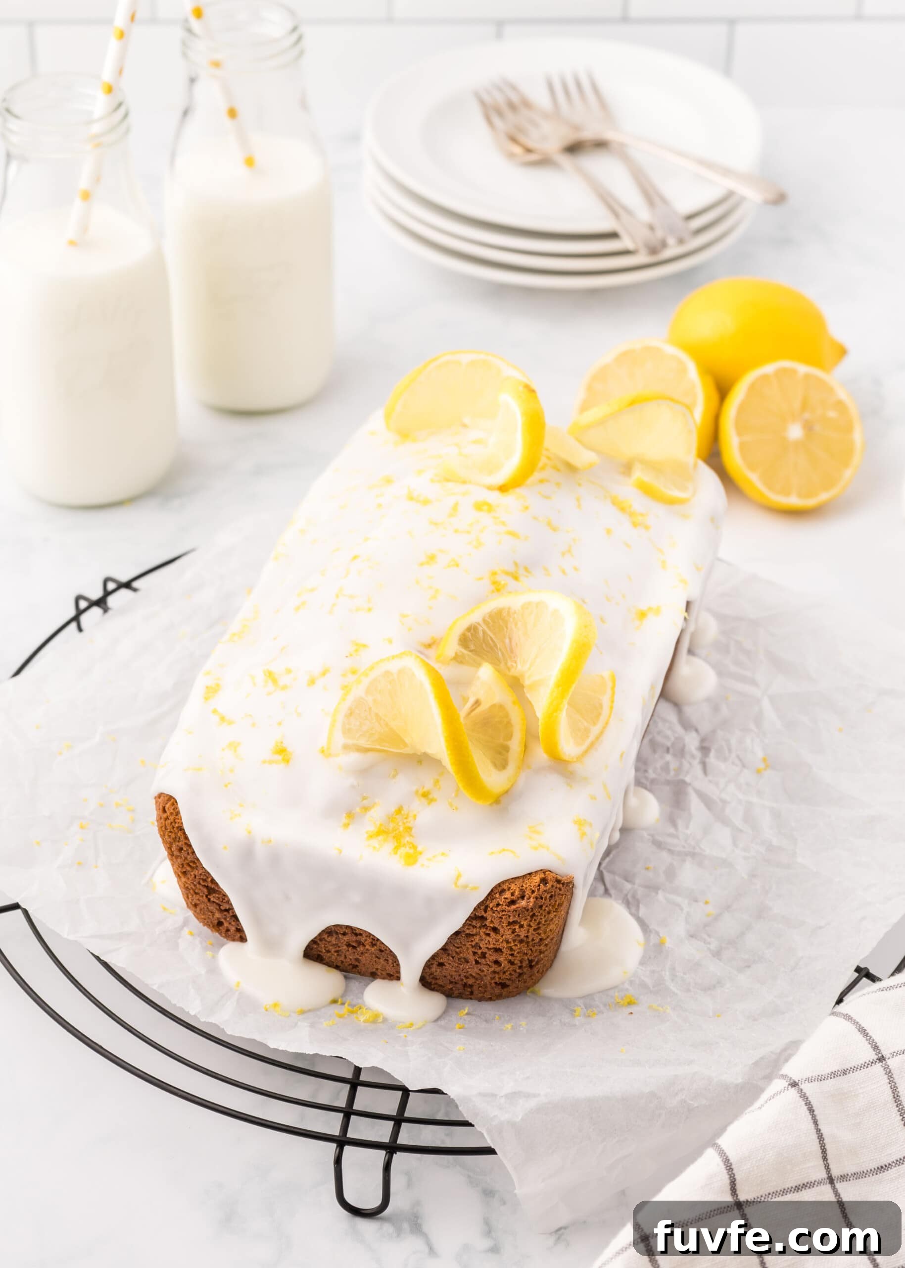 Zesty Iced Lemon Loaf Recipe for a Moist Citrus Cake 11 Lemon loaf with icing sprinkled with lemon zest and topped with lemon slices accented with glass milk bottles with white and yellow straws.