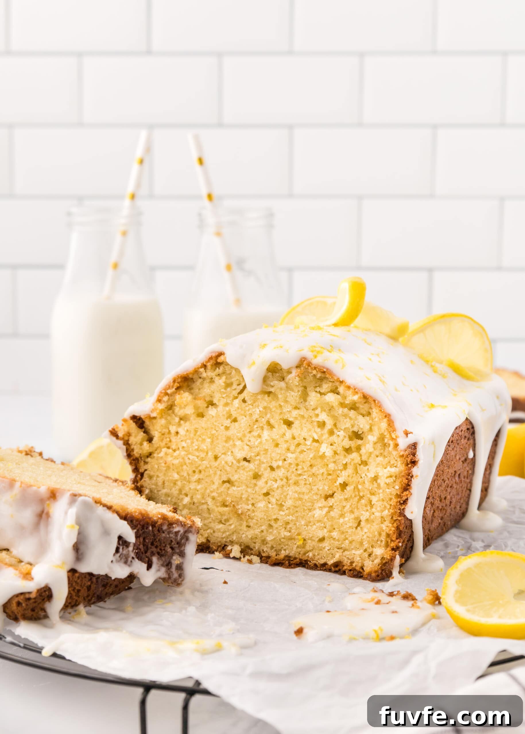 Zesty Iced Lemon Loaf Recipe for a Moist Citrus Cake 12 Lemon loaf with slices on a parchment paper lined cooling rack arranged in front of two glass bottles filled with milk.