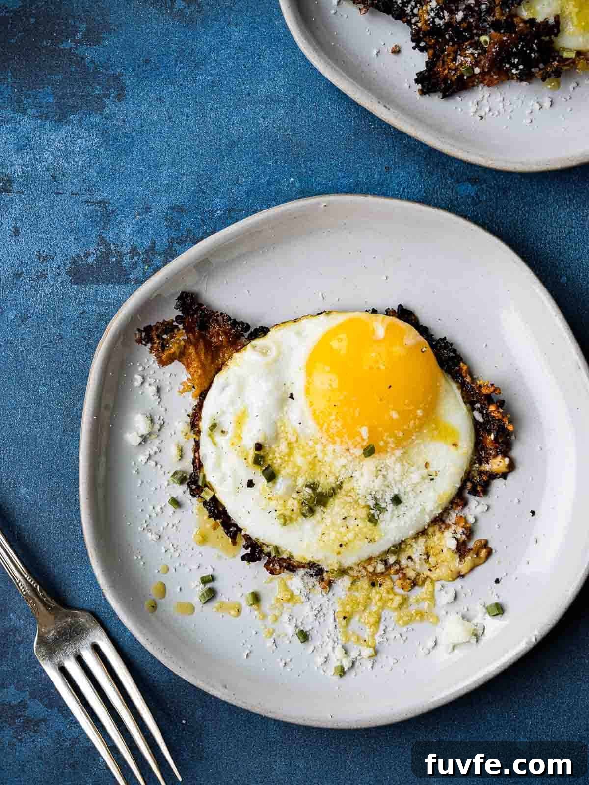 Parmesan Chili Crisp Fried Eggs: Crispy Sunny-Side with Heat 3 Overhead picture of a fried egg on a white plate, on top of a blue background.