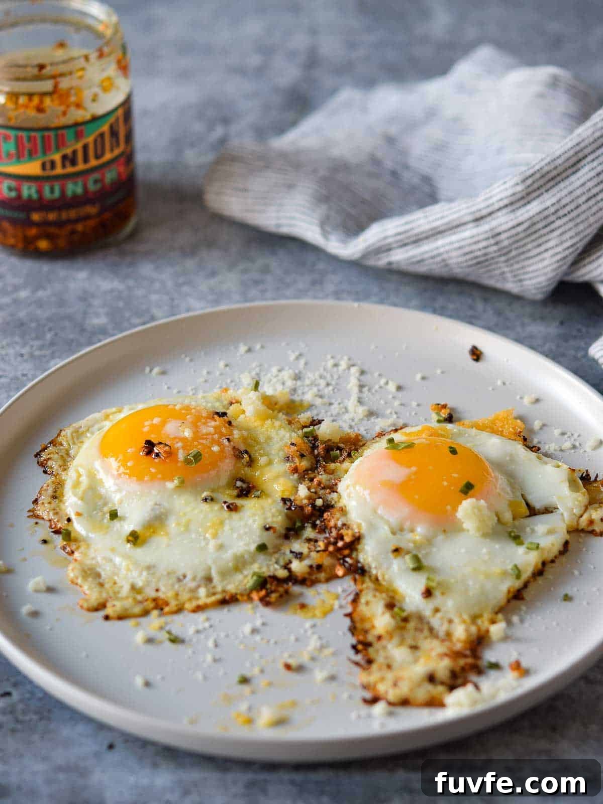 Parmesan Chili Crisp Fried Eggs: Crispy Sunny-Side with Heat 9 Angled view of two fried eggs on a plate with a jar of chili crunch in the background.