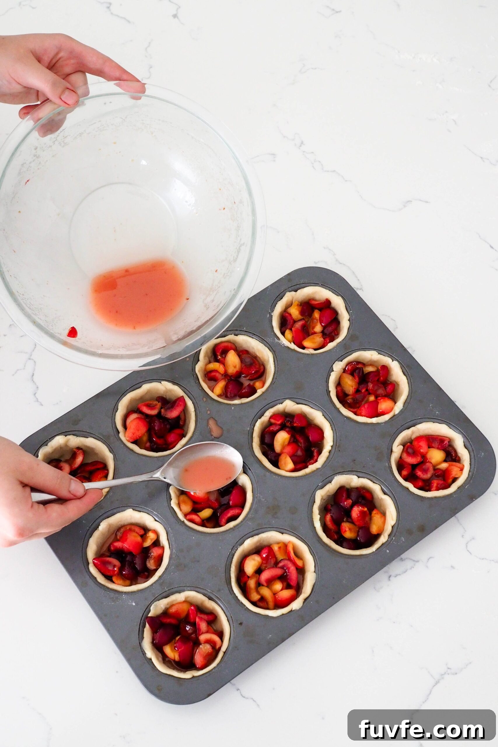 A hand uses a tablespoon to add pink liquid to mini cherry pies.