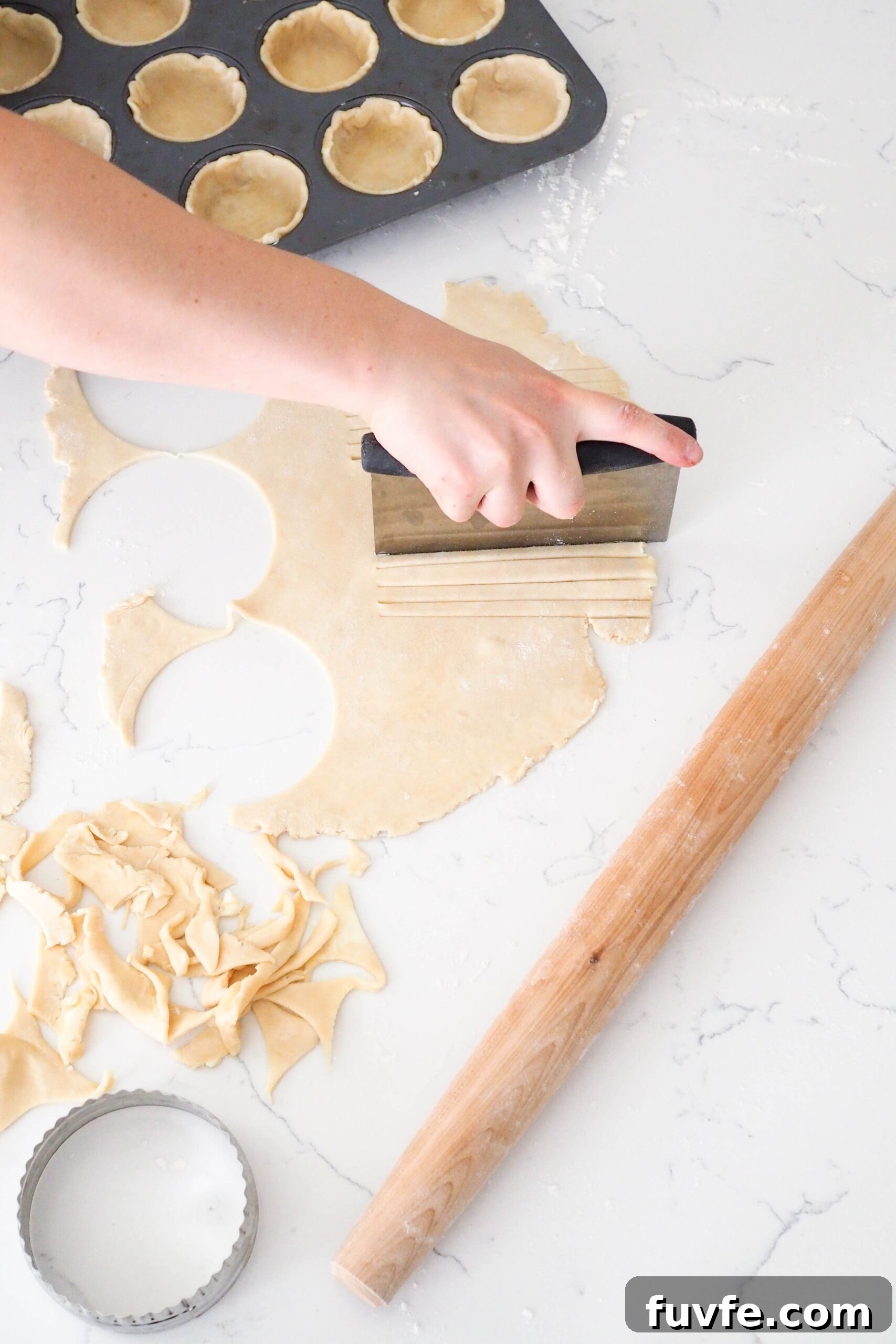 A hand uses a bench scraper to cut strips of pie dough.