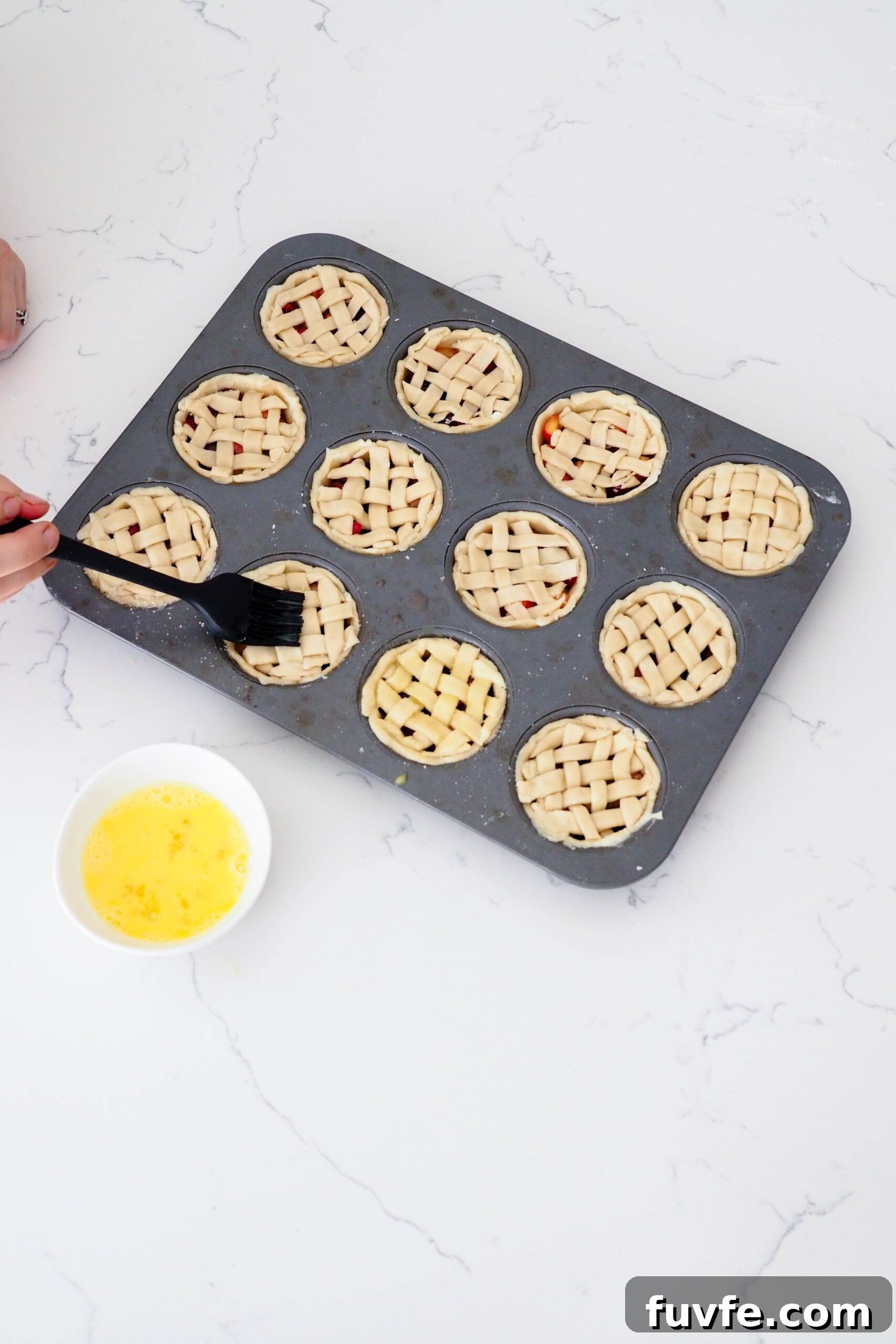 A hand uses a silicone pastry brush to brush egg wash onto mini latticed pies.