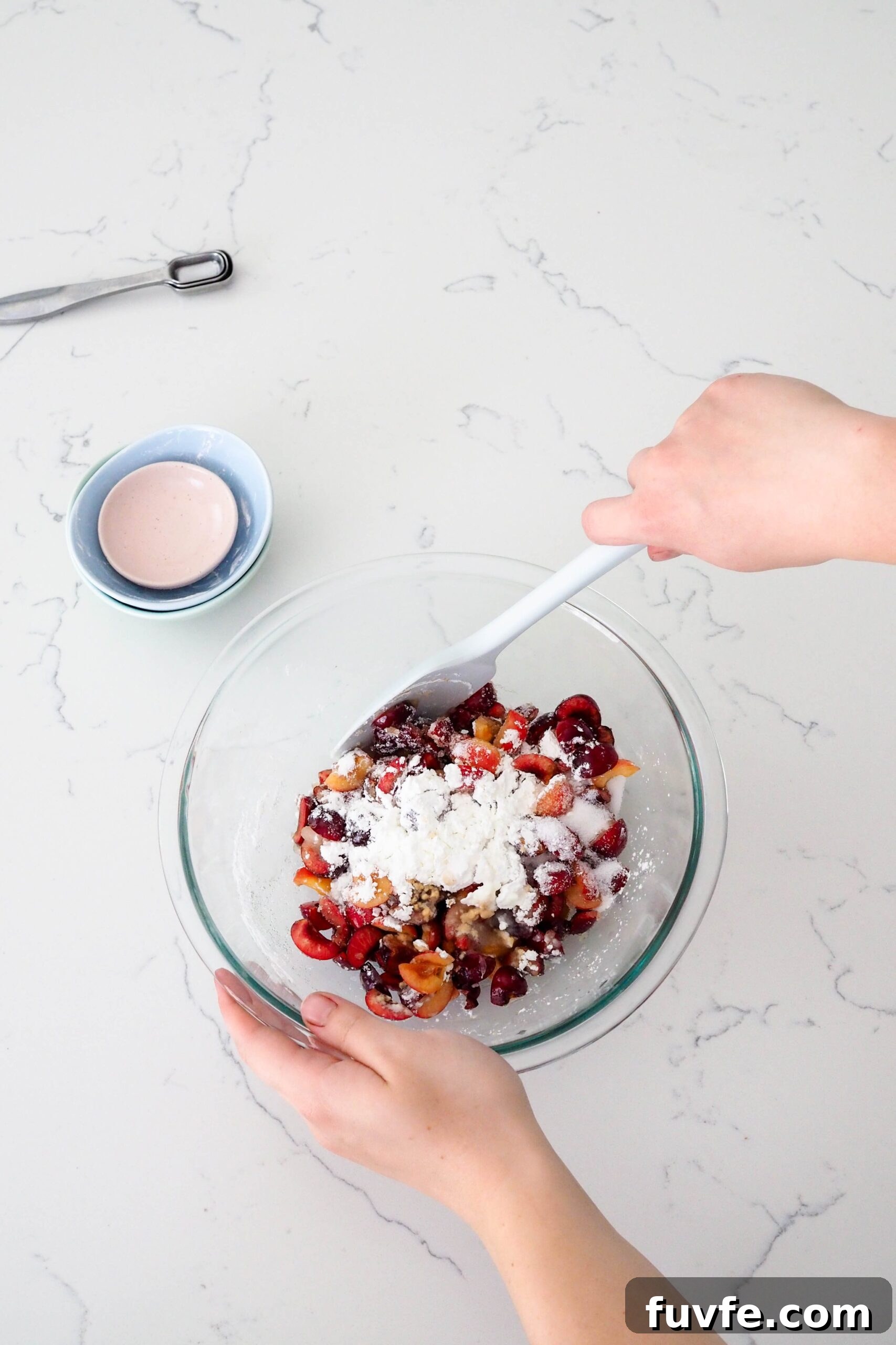 A hand stirs together cherries, cornstarch, and sugar in a bowl.