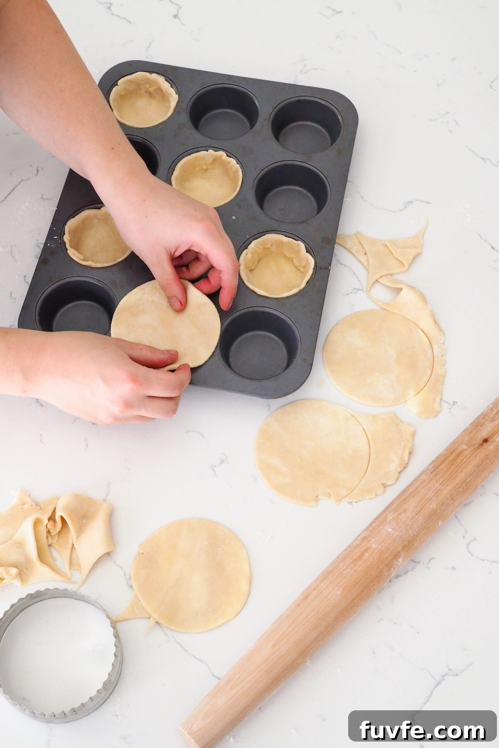 Two hands guide a circle of pie dough into a muffin pan opening.