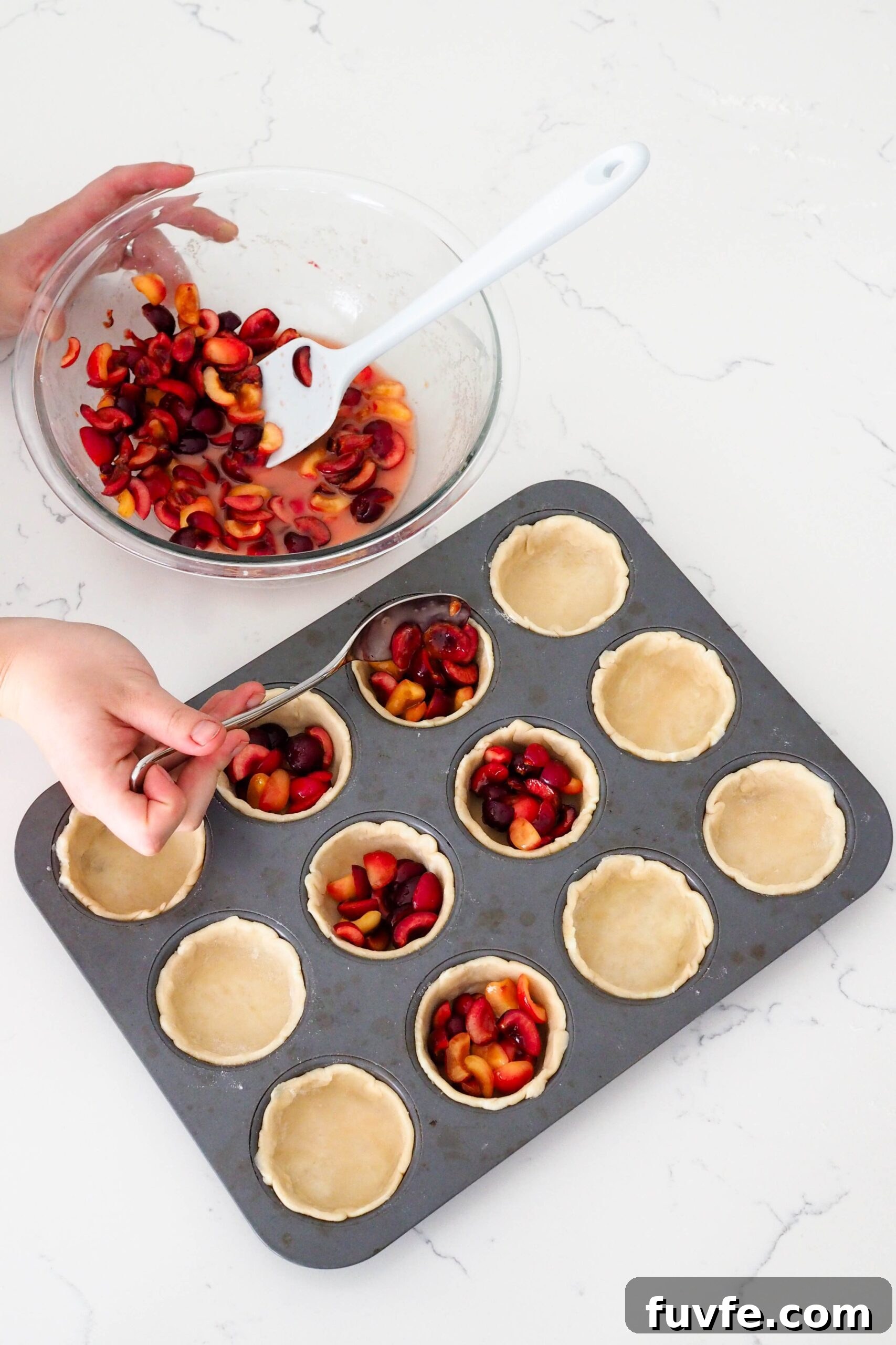 A hand uses a tablespoon to spoon cherries into mini pies.
