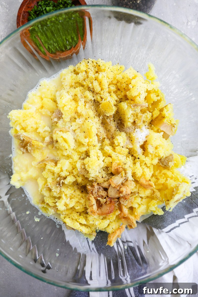 A large clear mixing bowl filled with mashed Yukon potatoes, butter, milk, roasted garlic, salt and pepper.
