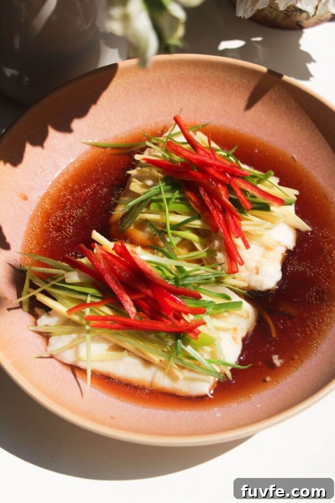 Soy sauce poured over fish fillets in pink bowl with scallions, chili and ginger batons on top.