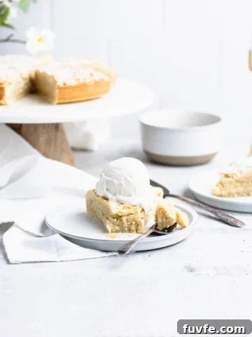 Slice of fyrstekake on a plate with a bite taken out of the center. In the background are a bouquet of white flowers, cake on a cake stand, a white bowl and a gold candle stick.