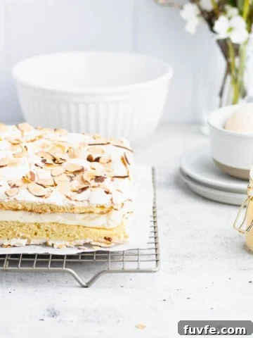 Verdens beste kake on a gold cooling rack with a white bowl, a jar of vaniljekrem, plates, eggs and a bouquet in the background.