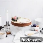 Frosted spice cake on a clear cake stand with while bowl, a jar of caramel, two taper candles and a small stack of white plates in the background.