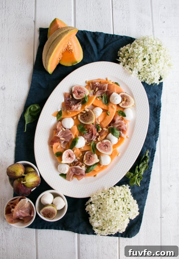 Overhead view of a melon prosciutto salad on a white platter. Individual ingredients surround the platter on a blue towel.