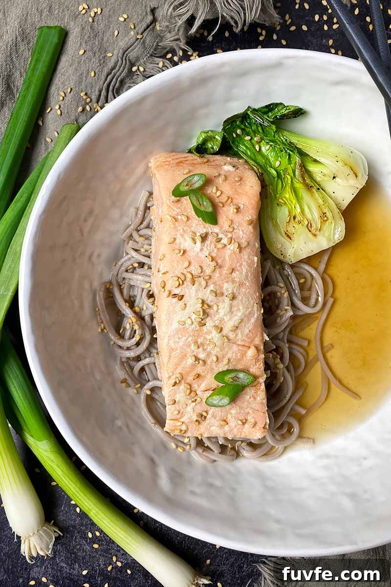 Vertical top-down image of a white bowl with broth, noodles, and a piece of fish.