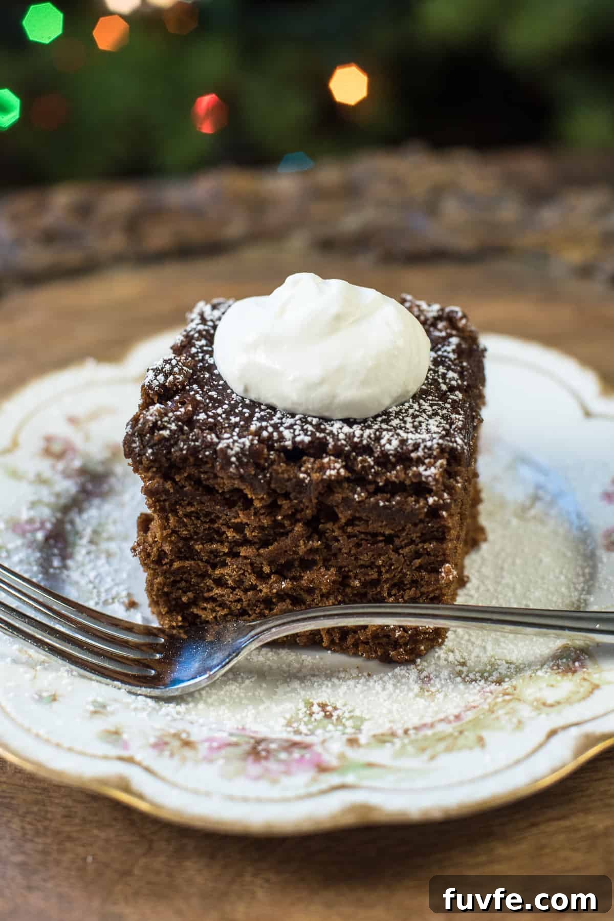 Warm Gingerbread Cake Recipe with Spiced Molasses Glaze 2 A slice of gingerbread topped with powdered sugar and whipped cream on a pretty china plate with a fork.