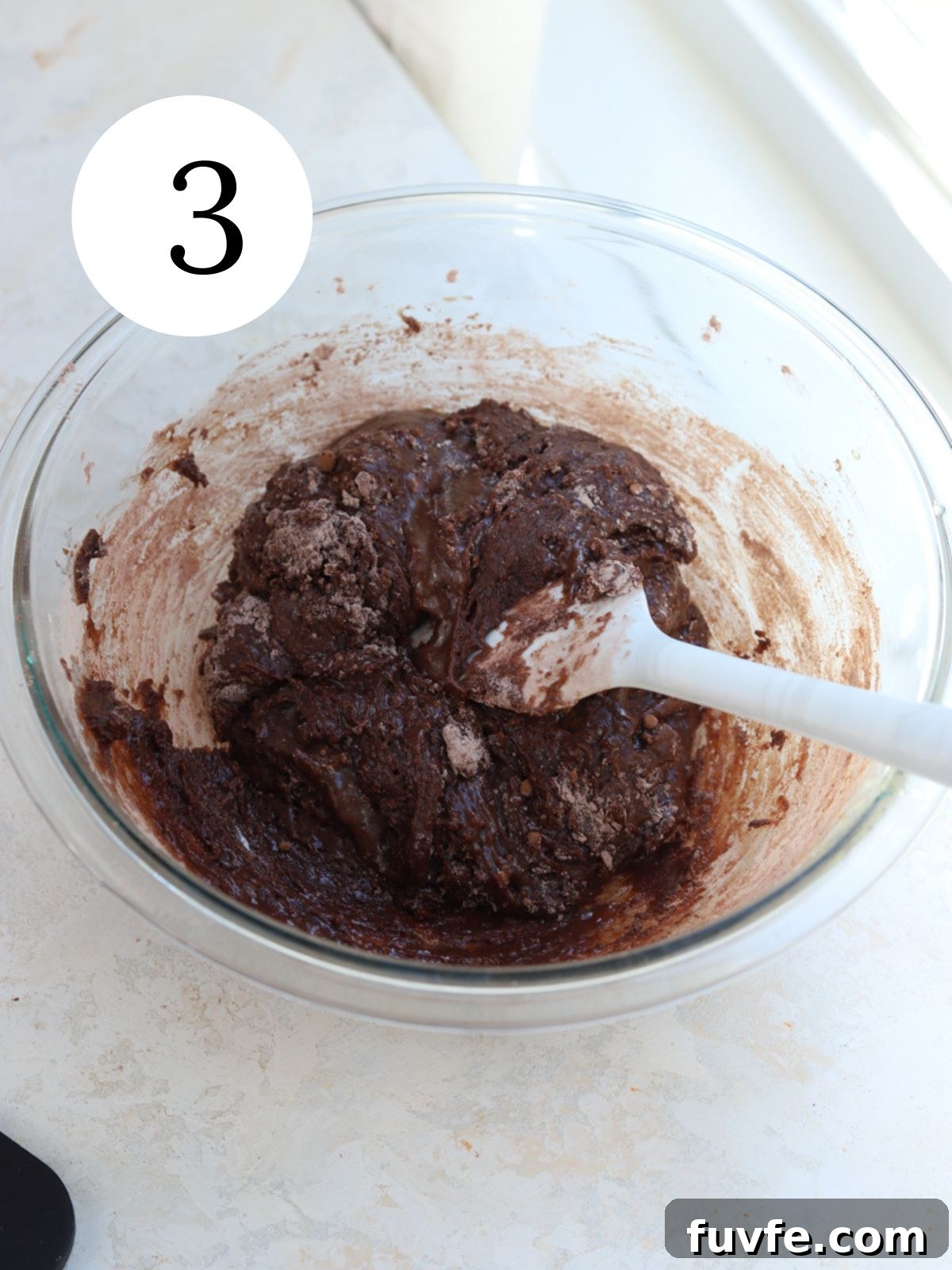 A glass mixing bowl with chocolate batter and a white spatula.