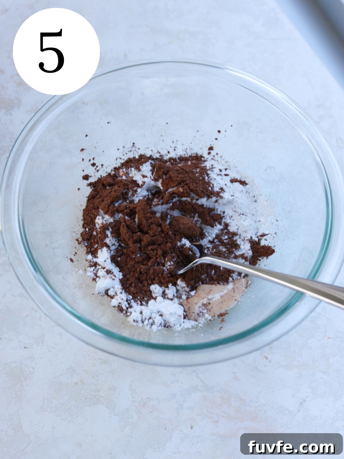 A glass mixing bowl with cocoa powder, powdered sugar and a spoon in it.
