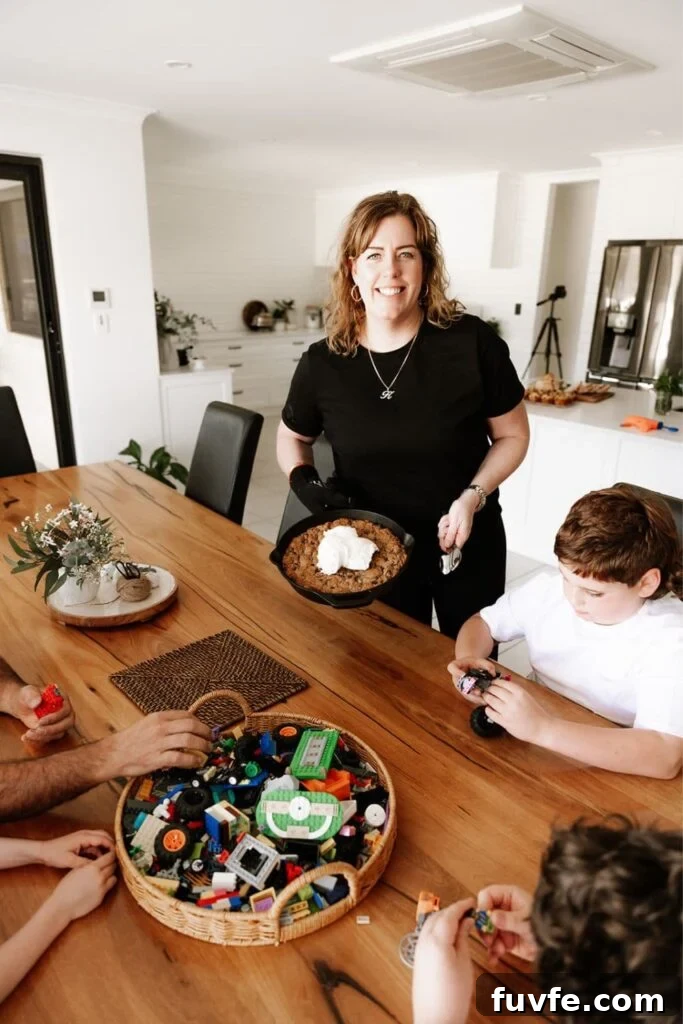 15 Irresistible Chocolate Sourdough Recipes to Try Today 27 Kate from The Pantry Mama holding a cast iron skillet filled with a chocolate chip sourdough cookie skillet. She is standing next to a dining room table where her family are playing Lego.