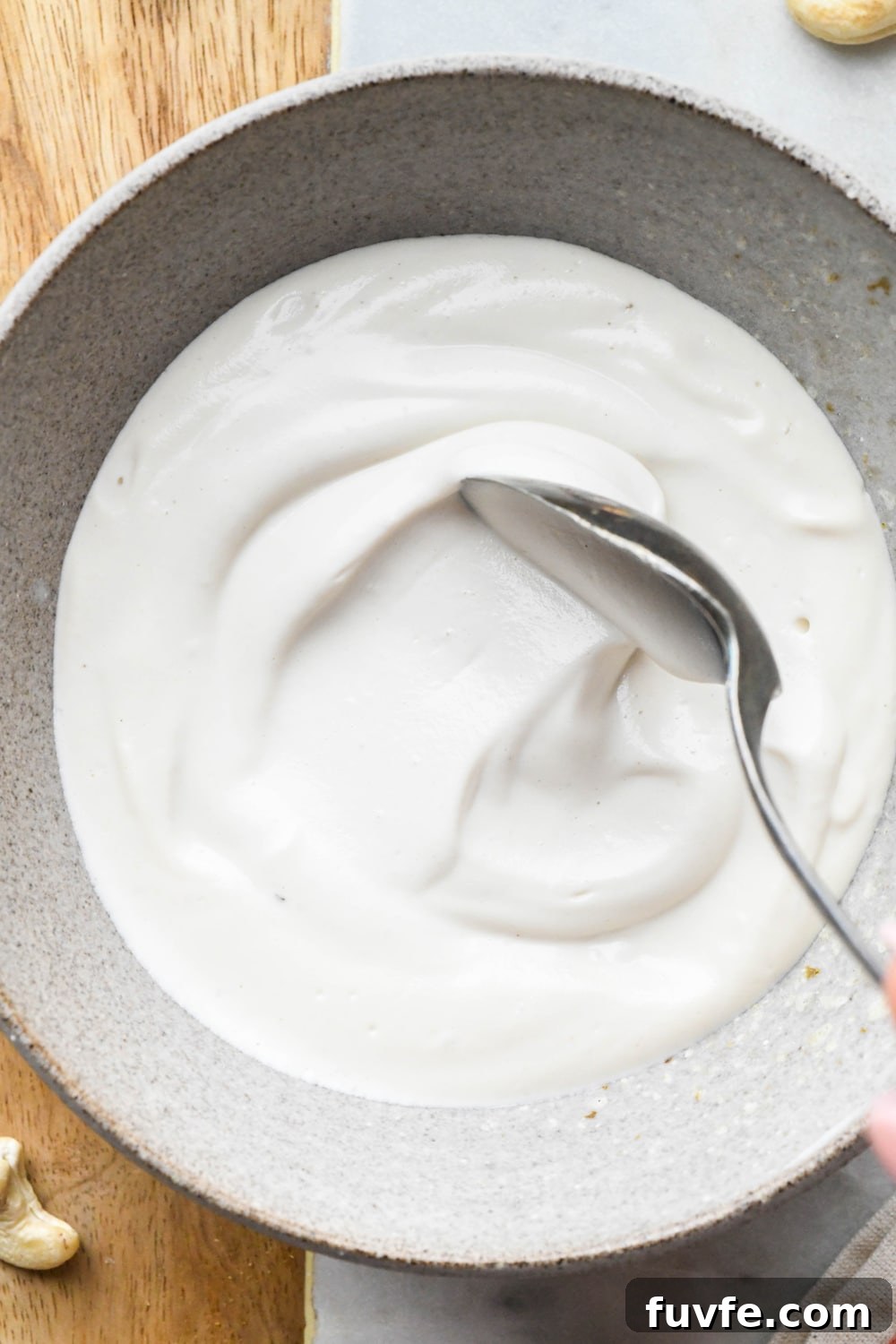 Cashew cream made with 1/2 cup of water in a grey ceramic bowl with a spoon stirring the cream to show the texture.