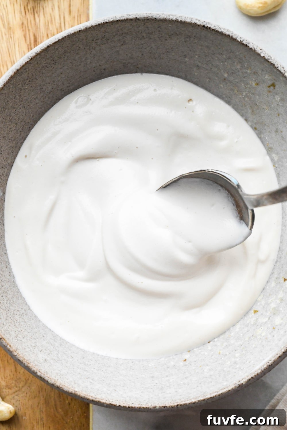 Cashew cream made with 1/2 cup of water in a grey ceramic bowl with a spoon lifting out some of the cream to show the texture.