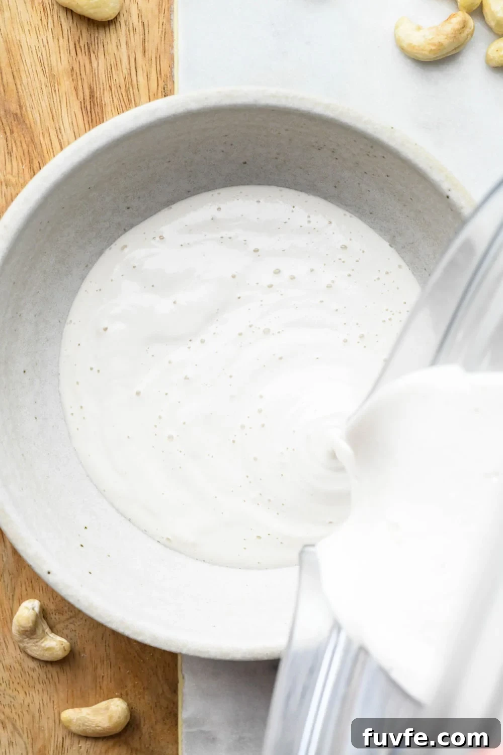 A blender pouring cashew cream made with 3/4 cup of water into a grey ceramic bowl. 
