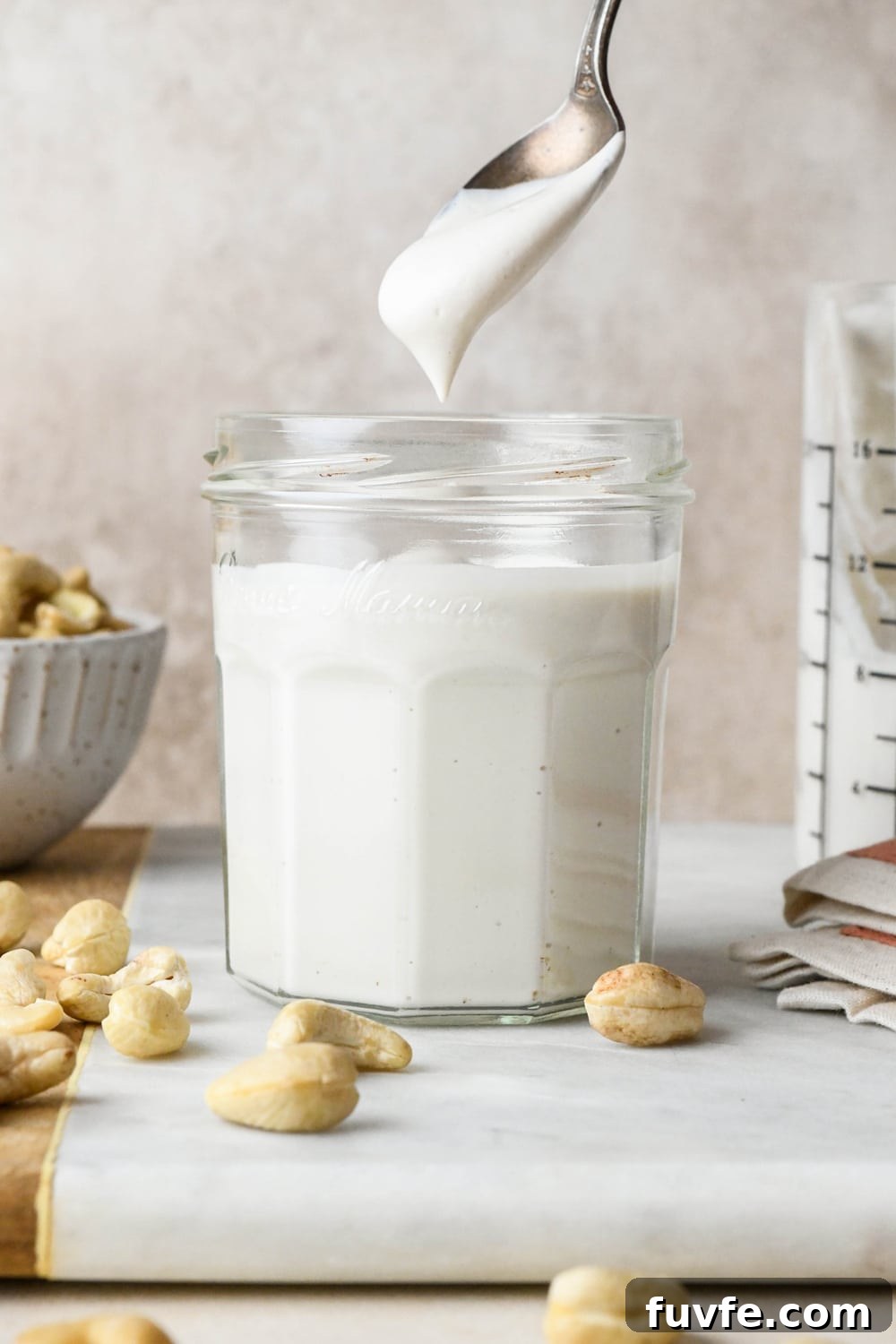 A hand lifting out a spoonful of cashew cream from a small jar surrounded by a dish and some scattered raw cashews.