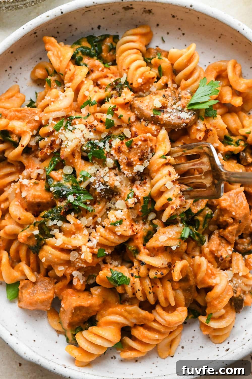 Creamy tomato sausage pasta in a shallow white speckled bowl, topped with dairy free parmesan, chili flakes, and a fresh herbs, with a fork lifting out a bite of pasta.