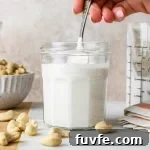 A hand lifting out a spoonful of cashew cream from a small jar surrounded by a dish and some scattered raw cashews.