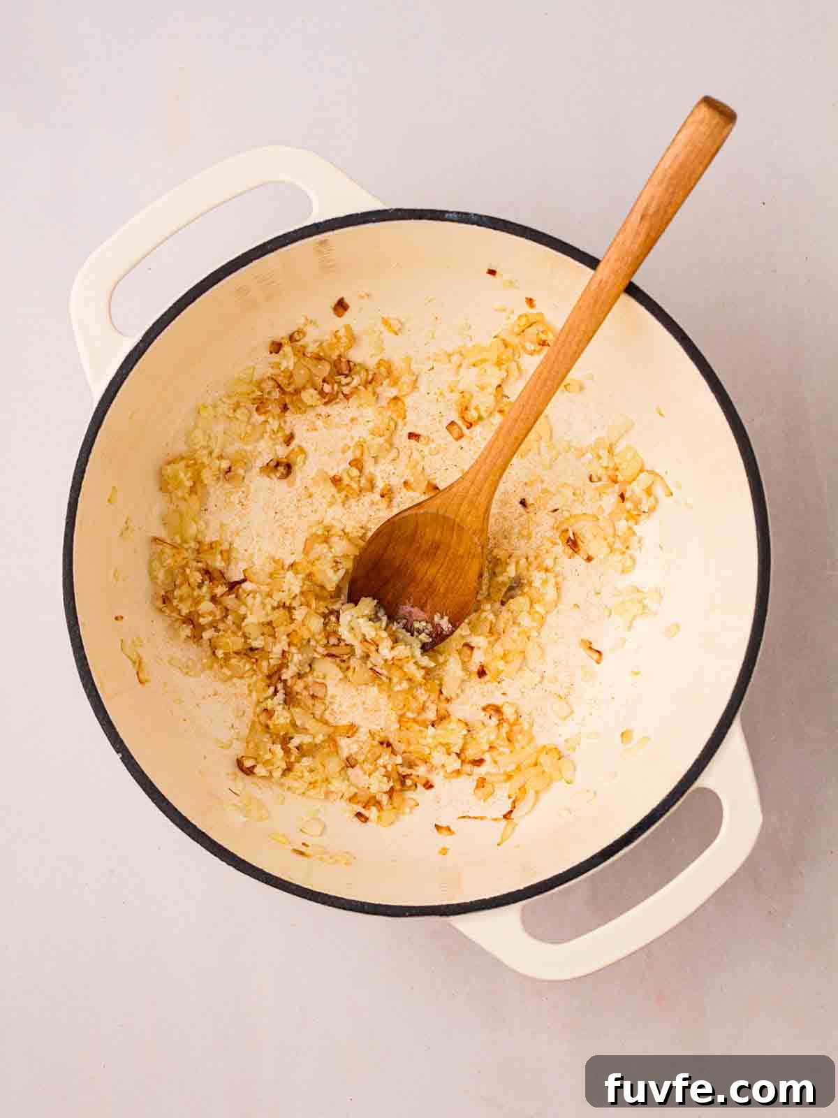Onions being sautéed in a large pan with a wooden spoon in.