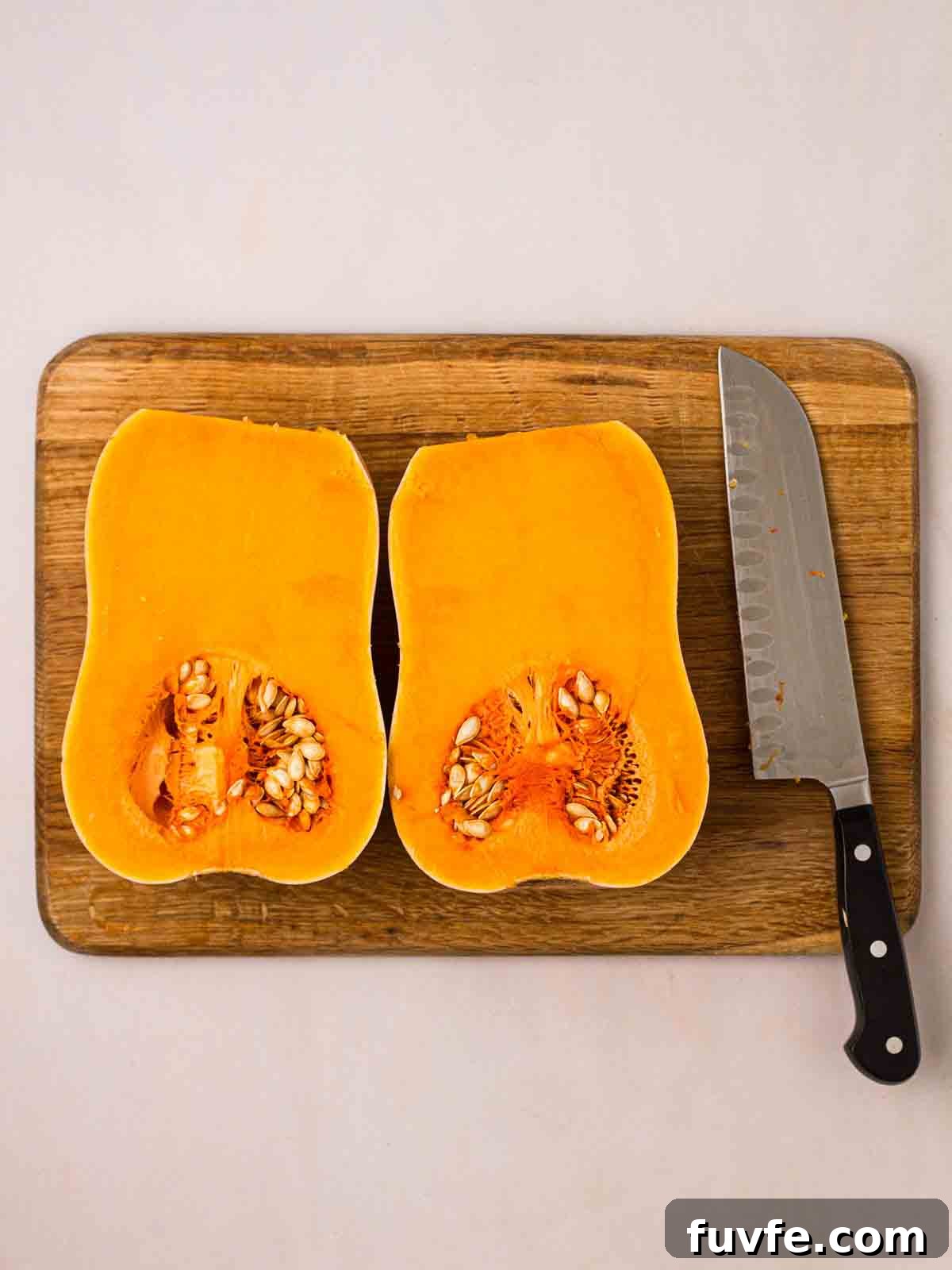 A halved butternut squash on a chopping board with a knife to the side.
