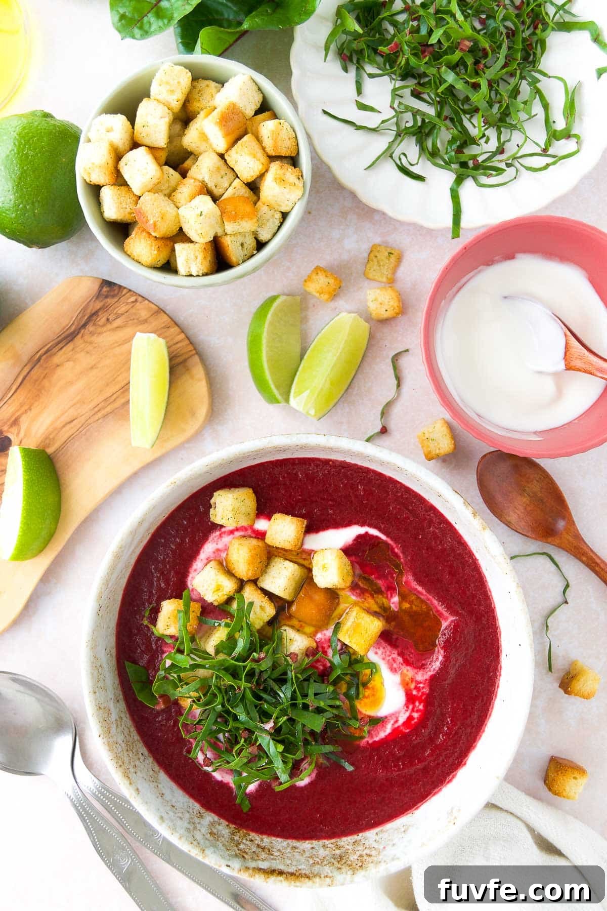 Cold beet soup in a white bowl topped with Greek yogurt swirl, croutons, and beet greens, with lime wedges and yogurt on the side