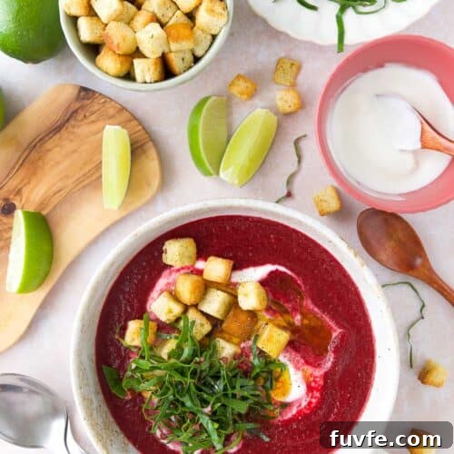 Cold beet soup in a white bowl topped with Greek yogurt swirl, croutons, and beet greens, with lime wedges and yogurt on the side